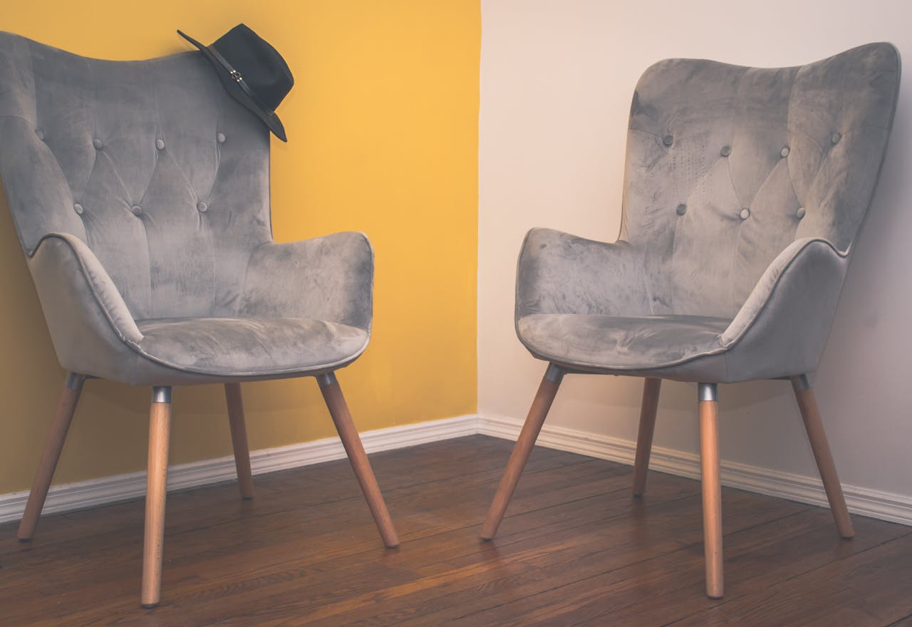 services-05 Two contemporary gray armchairs in a minimalist interior with a yellow accent wall and wooden flooring.